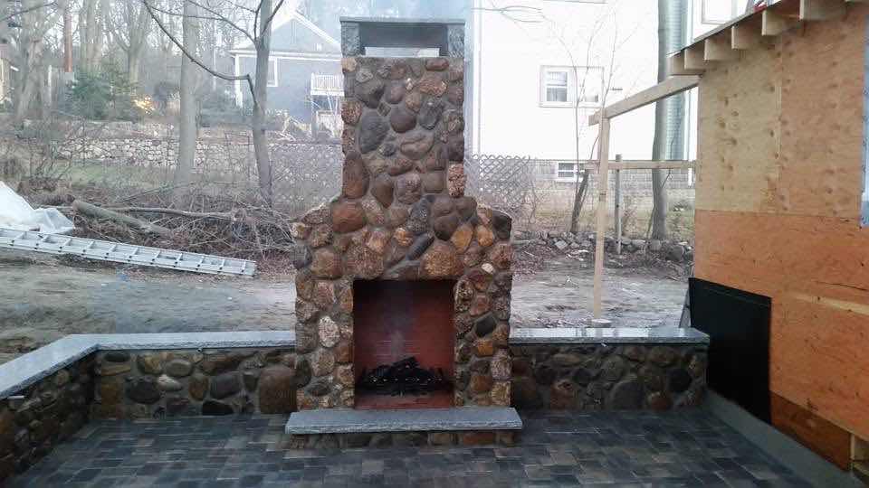 Fieldstone outdoor fireplace with a lit fire at dusk, built by Magic Tools Contracting